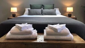 Una cama bien hecha con edredón gris y almohadas, dos lámparas de noche y dos pilas de toallas dobladas sobre una mesa de madera.