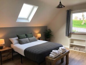 Ein Schlafzimmer mit Oberlicht, einem Bett mit Kissen, Nachttischlampen, einer Pflanze, einem Fenster mit Vorhängen und einer Holzbank mit Handtüchern.
