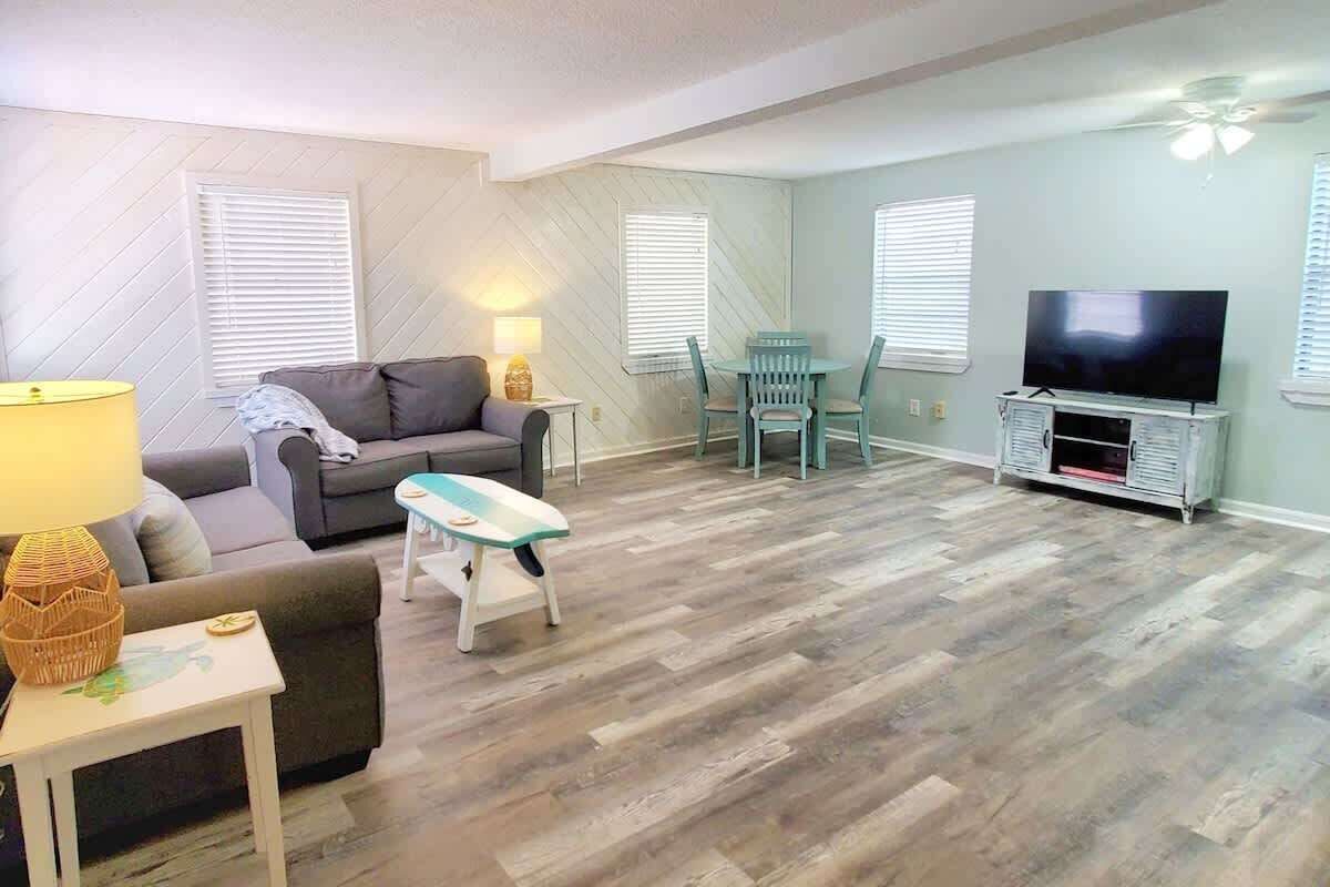 Un salón con un sofá, una mesita de noche con una lámpara, un mueble de televisión con televisor, y un comedor con sillas y una mesa.