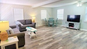 Un salón con un sofá, una mesita de noche con una lámpara, un mueble de televisión con televisor, y un comedor con sillas y una mesa.