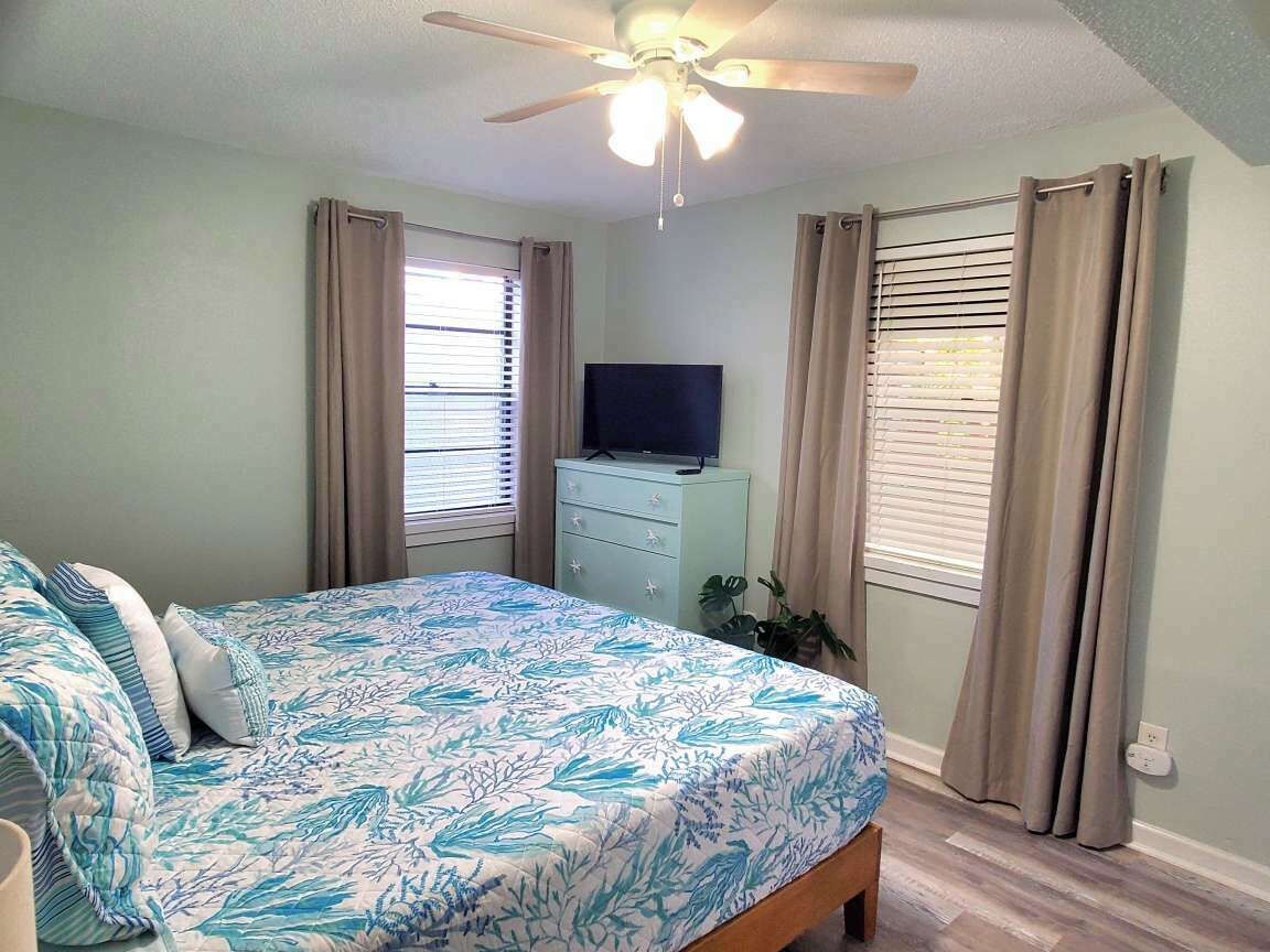 A bedroom with a bed, a ceiling fan, a TV on a dresser, and windows with blinds.