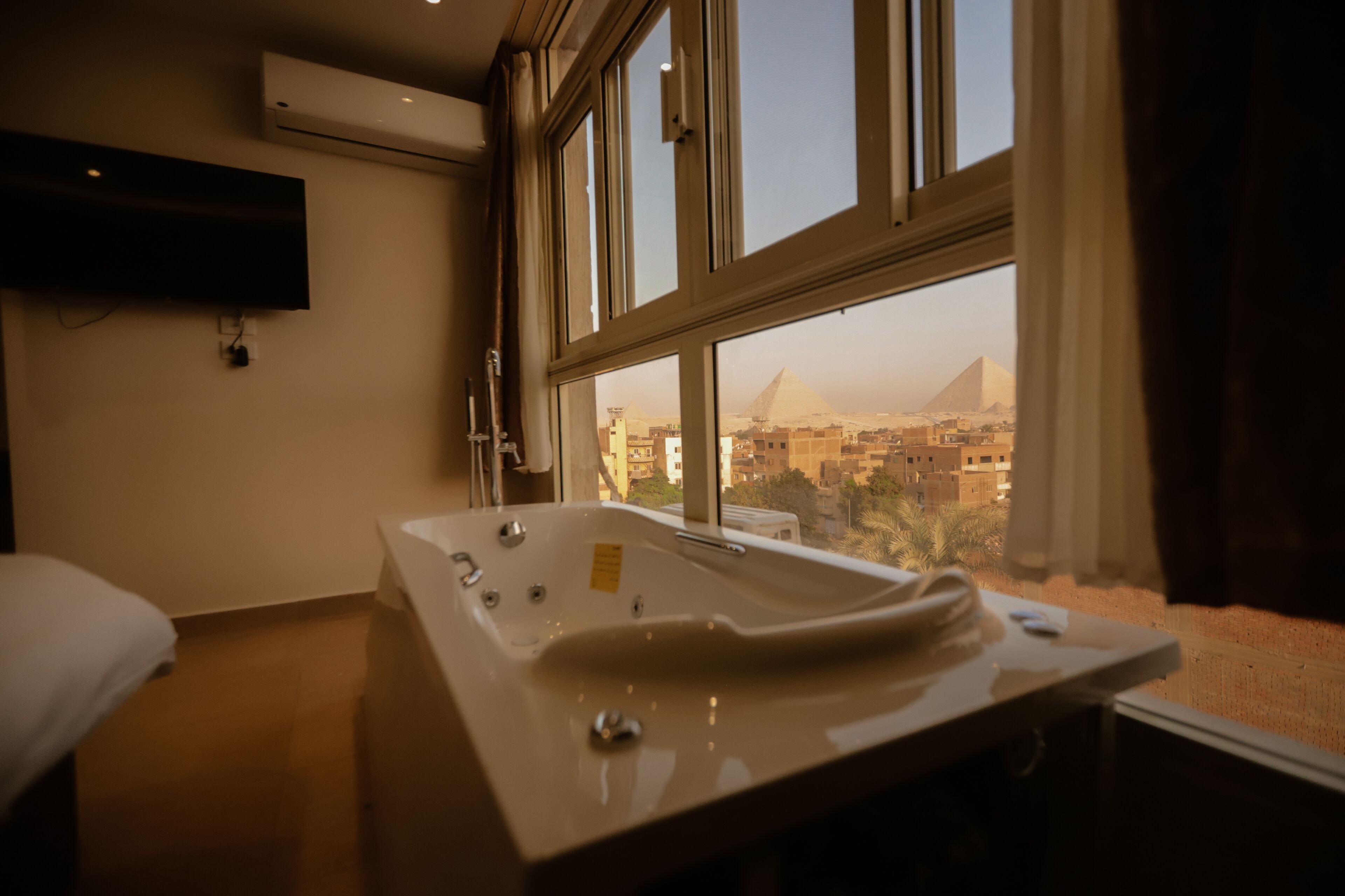A hotel room with a large bathtub, a flat-screen TV mounted on the wall, and a view of a pyramid through the window.