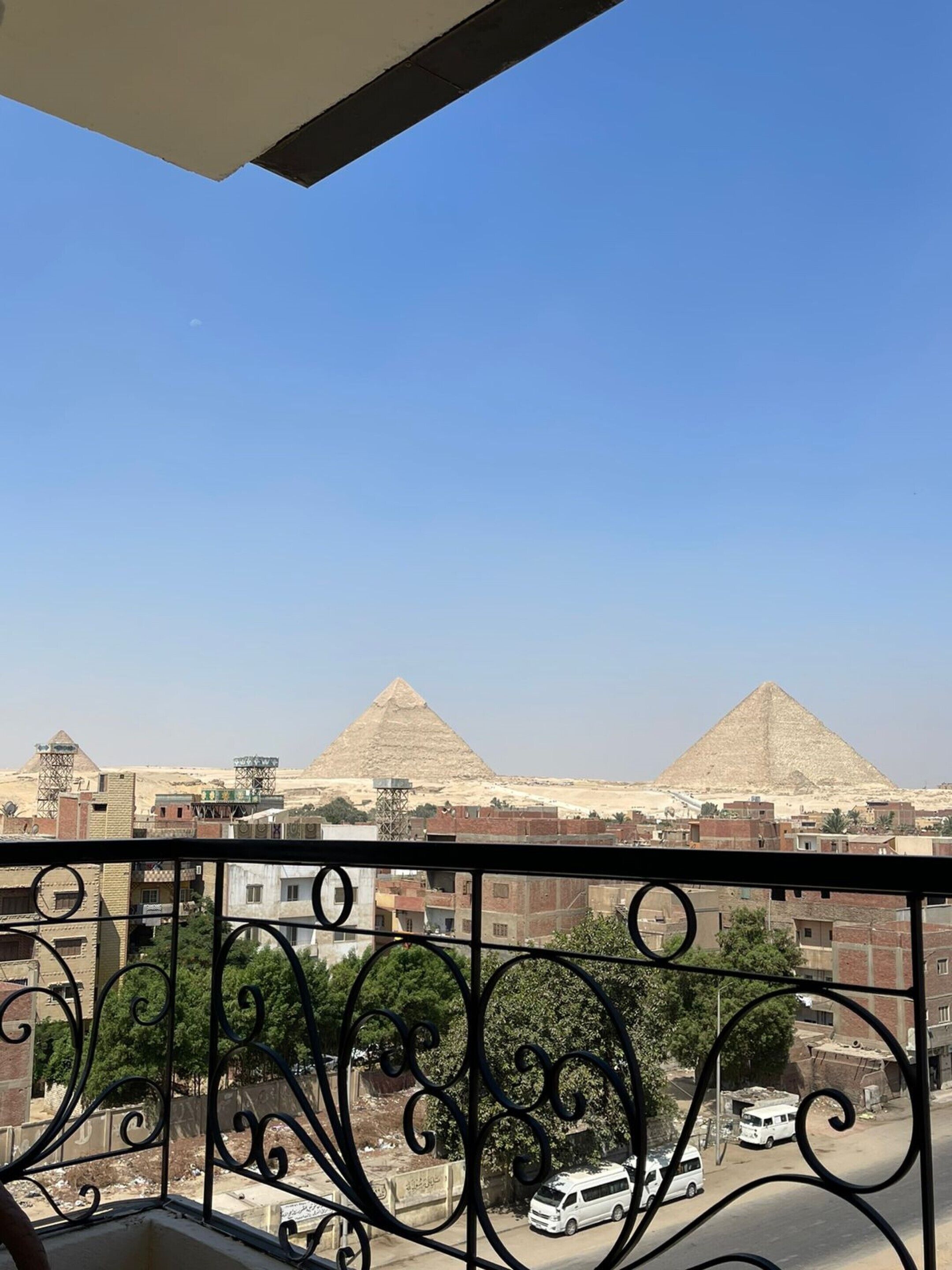 A balcony with a view of two pyramids in the distance, a clear blue sky, and a street with parked cars.