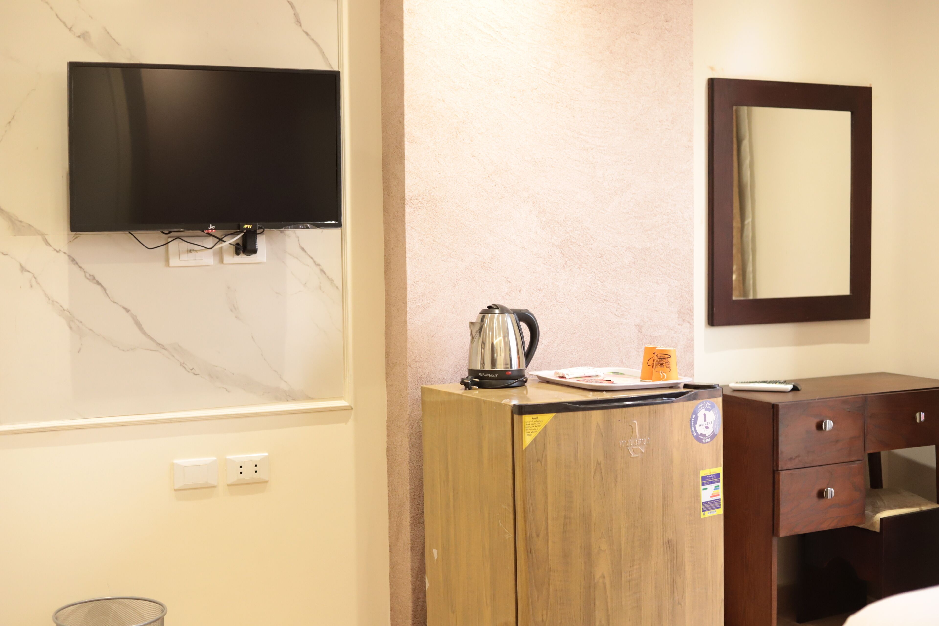 A hotel room with a flat-screen TV mounted on the wall, a wooden cabinet with a kettle, and a small table with a cup.