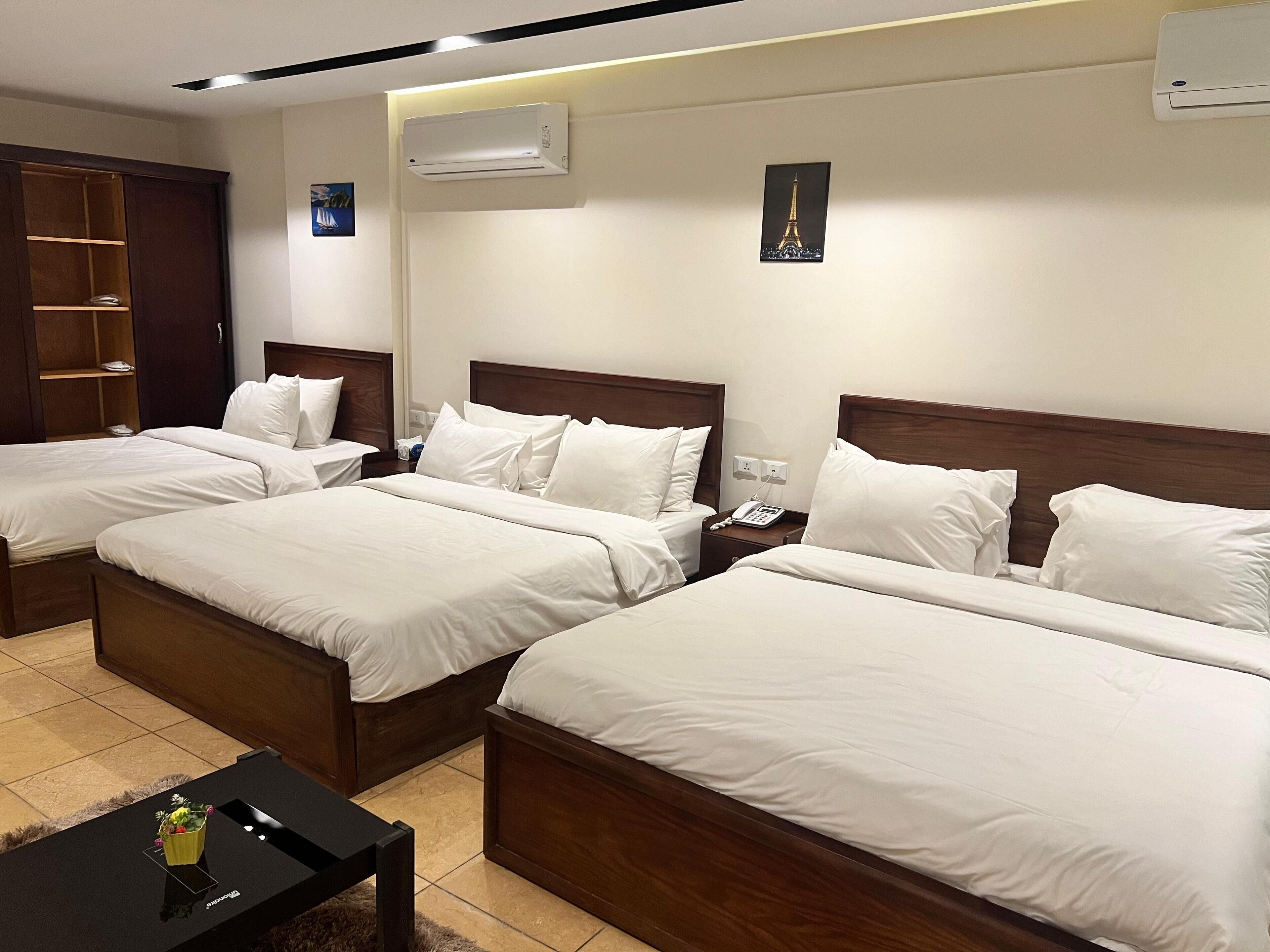 A hotel room with three single beds, a wooden wardrobe, a bedside table with a phone, and a small table with a vase.