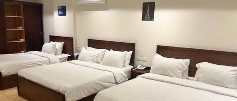 A hotel room with three single beds, a wooden wardrobe, a bedside table with a phone, and a small table with a vase.