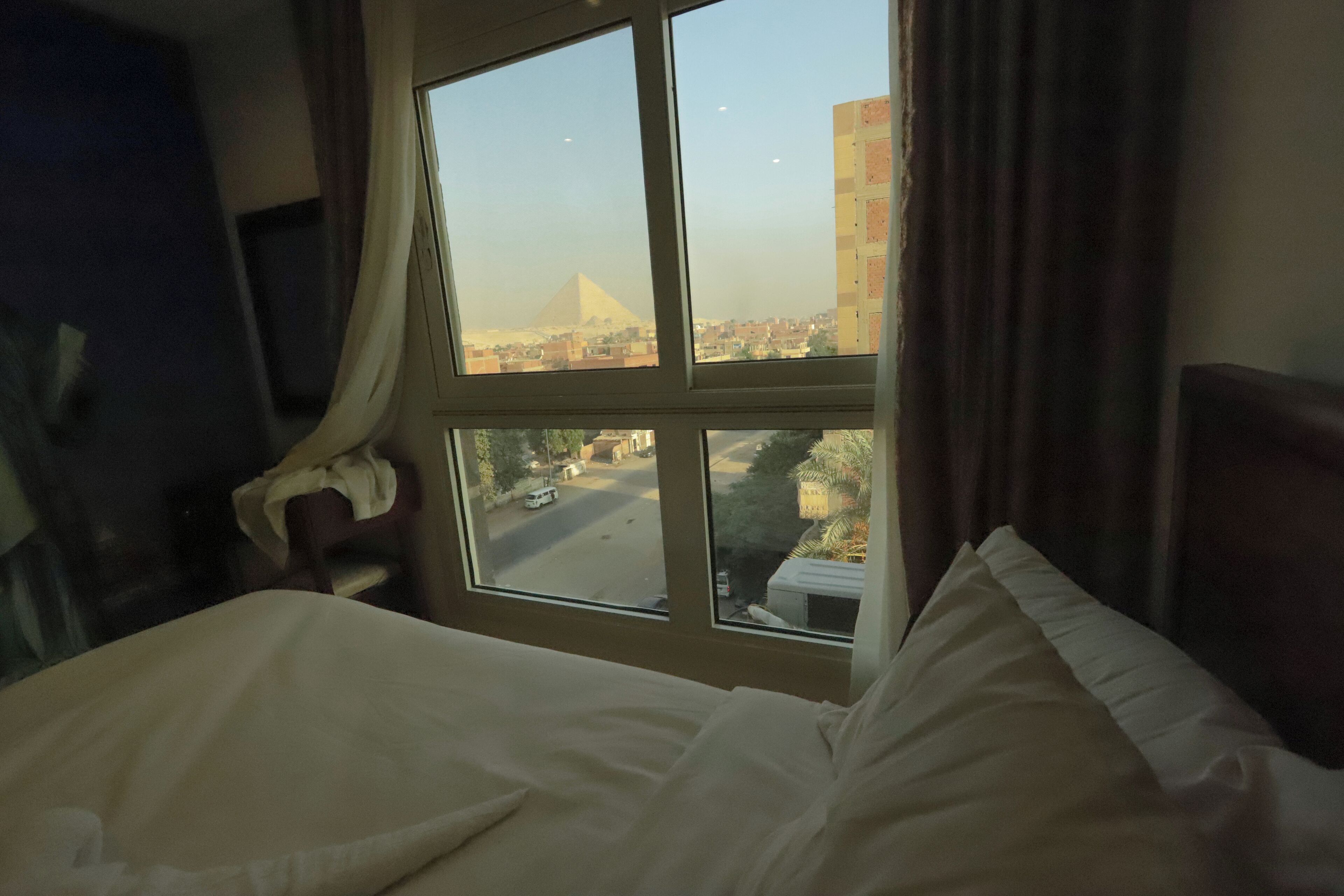 A hotel room with a bed, a window with a view of a pyramid and cityscape.