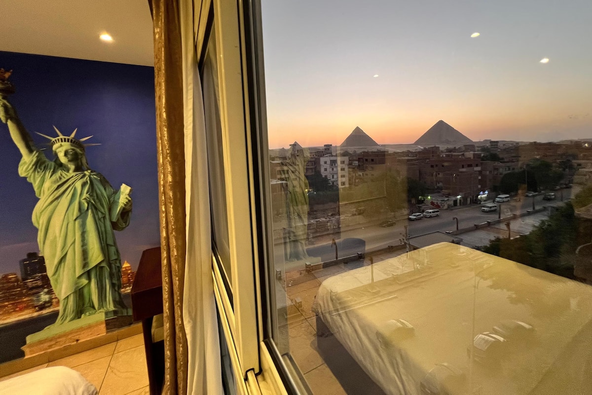 Huone, josta avautuu näkymä pyramideille auringonlaskun aikaan, Vapaudenpatsaan patsas ja valkoisella päällisellä varustettu sänky.