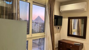 A hotel room with a large window, an air conditioning unit, a flat-screen TV, a wooden cabinet, and a mirror. - King Khafren View INN (Giza)