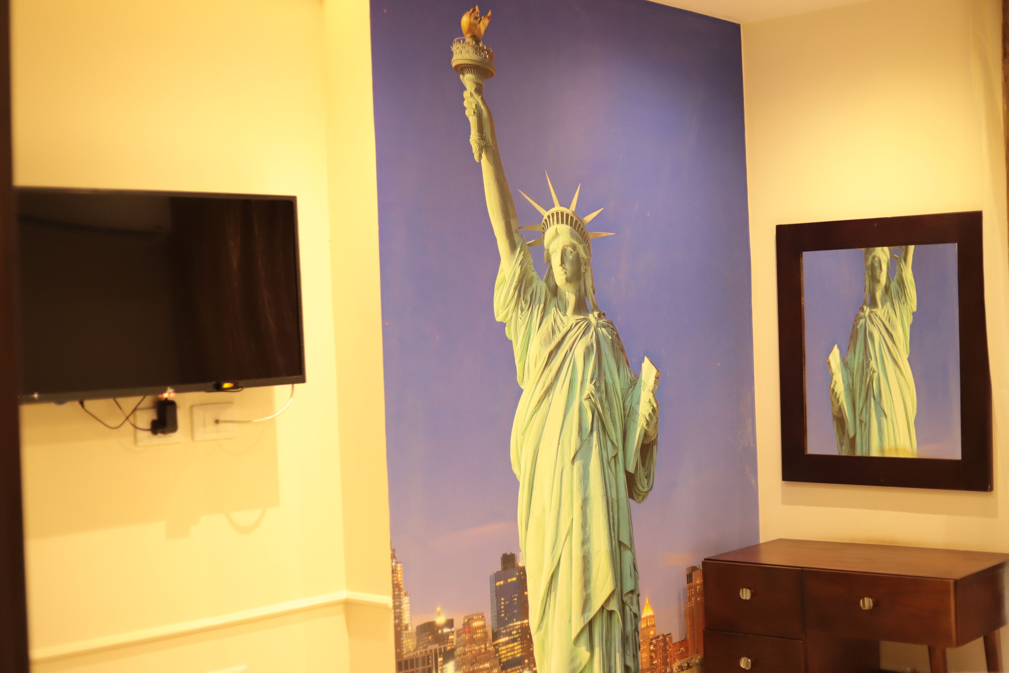 A room with a large wall mural of the Statue of Liberty, a flat-screen TV mounted on the wall, and a wooden dresser.