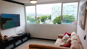 A living room with a beige sofa, a flat-screen TV mounted on the wall, a black entertainment unit, and a large window with a view of buildings and trees. - Cozy Elegance : Tu Rincón en San Juan (San Juan)