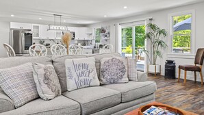 A modern living room with a grey sofa, decorative pillows, a coffee table, and a view of the kitchen and outside.