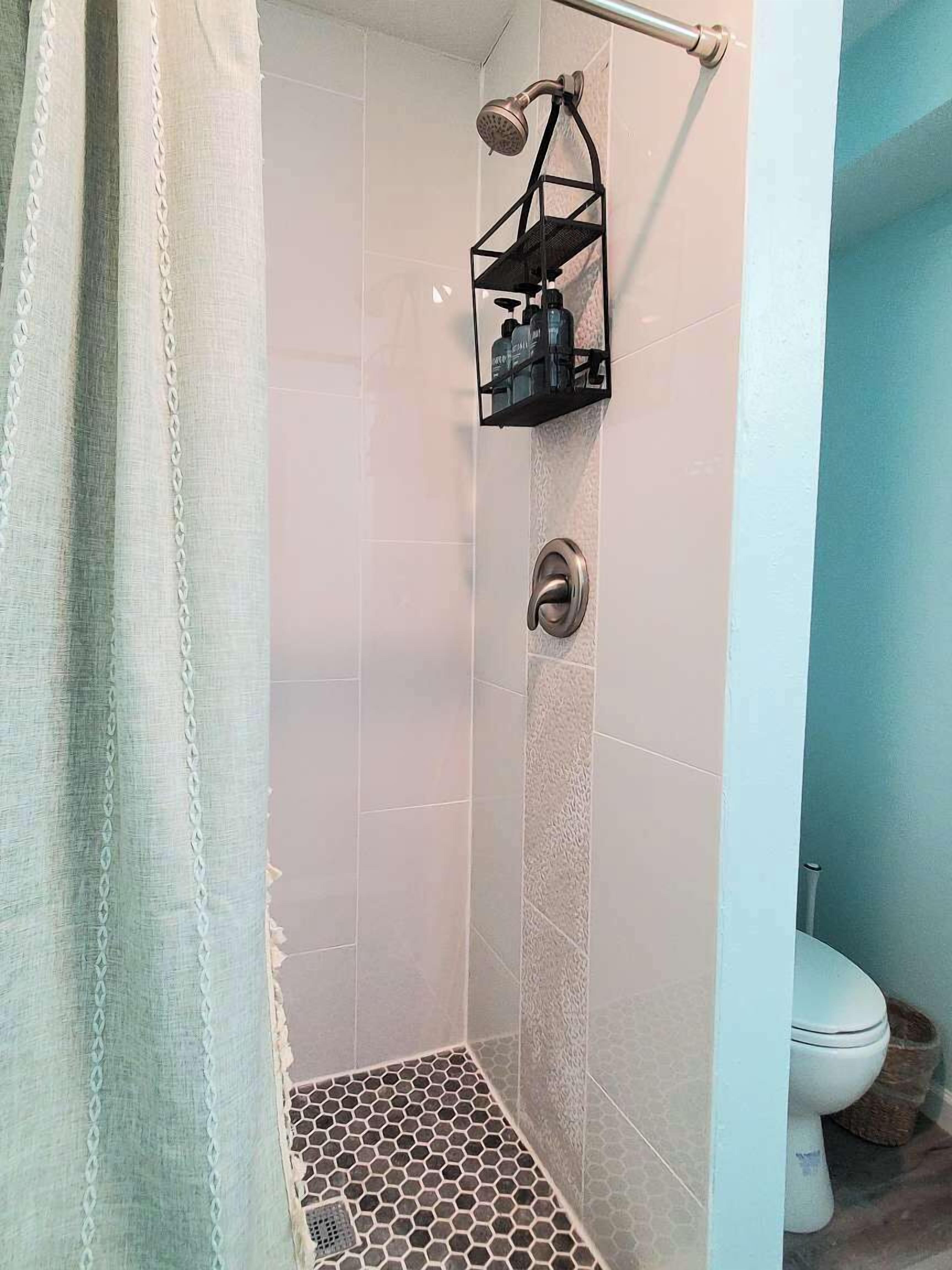 A shower with a hexagonal tile floor, a metal showerhead, and a black shelf with toiletries.