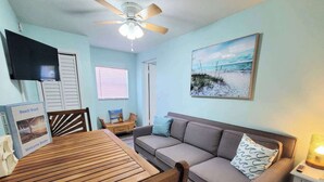 A room with a wooden table, a sofa, a ceiling fan, a TV, and a beach-themed painting.