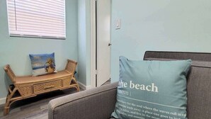 A room with a light blue wall, a wooden bench with cushions, a grey sofa, and a pillow with text.