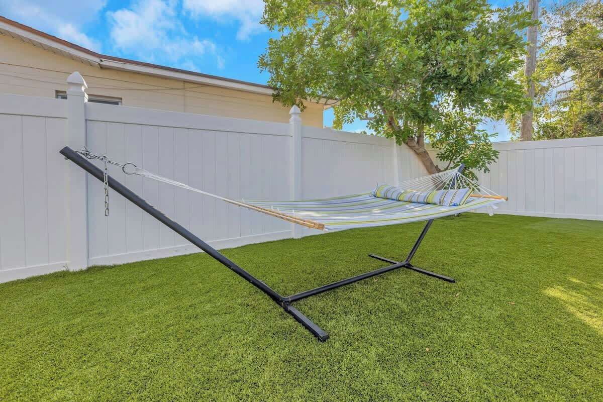 A hammock set up on a grassy lawn with a white fence and a tree in the background.