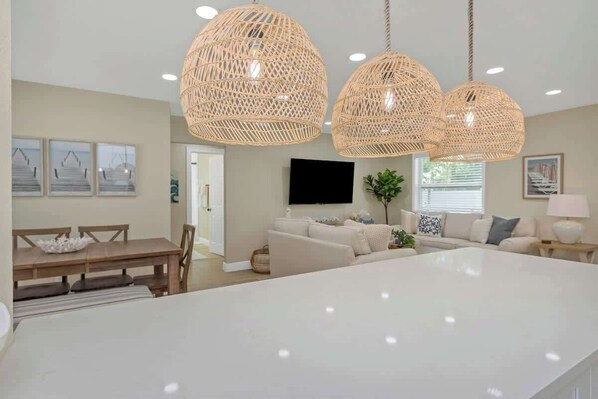 A modern kitchen with a large island, dining area with wooden table and chairs, and a living room with a sofa and TV.