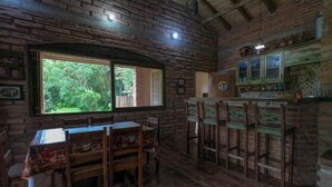 A rustic wooden interior with a dining table and chairs, a kitchen area with a bar, and a large window offering a view of greenery. - RECANTO DOS COMPADRES (Praia Grande)