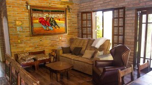 A cozy room with a brick wall, a sofa, a chair, and a wooden table. - RECANTO DOS COMPADRES (Praia Grande)