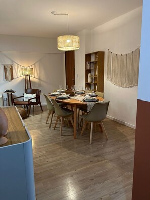 A dining area with a wooden table set for six, surrounded by chairs. There is a bookshelf with books and decorative items, a macrame wall hanging, and a floor lamp.