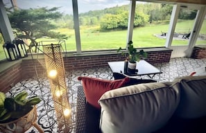 Une pièce chaleureuse avec une grande fenêtre donnant sur un espace verdoyant. On y trouve un fauteuil rembourré de couleur rouge, une petite table sur laquelle est posée une plante en pot, et un luminaire décoratif.
