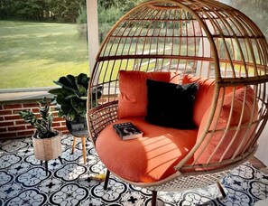 Un fauteuil suspendu en osier, doté de coussins orange et d’un oreiller noir, est placé sur un sol à motifs, offrant une vue sur une zone herbeuse à travers une fenêtre.