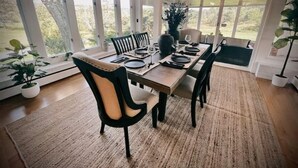 A dining area with a long table set for four, surrounded by large windows offering a view of greenery.