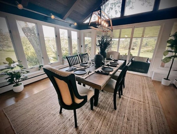 Un espace repas avec une longue table dressée pour quatre personnes, entouré de grandes fenêtres donnant sur un paysage verdoyant.