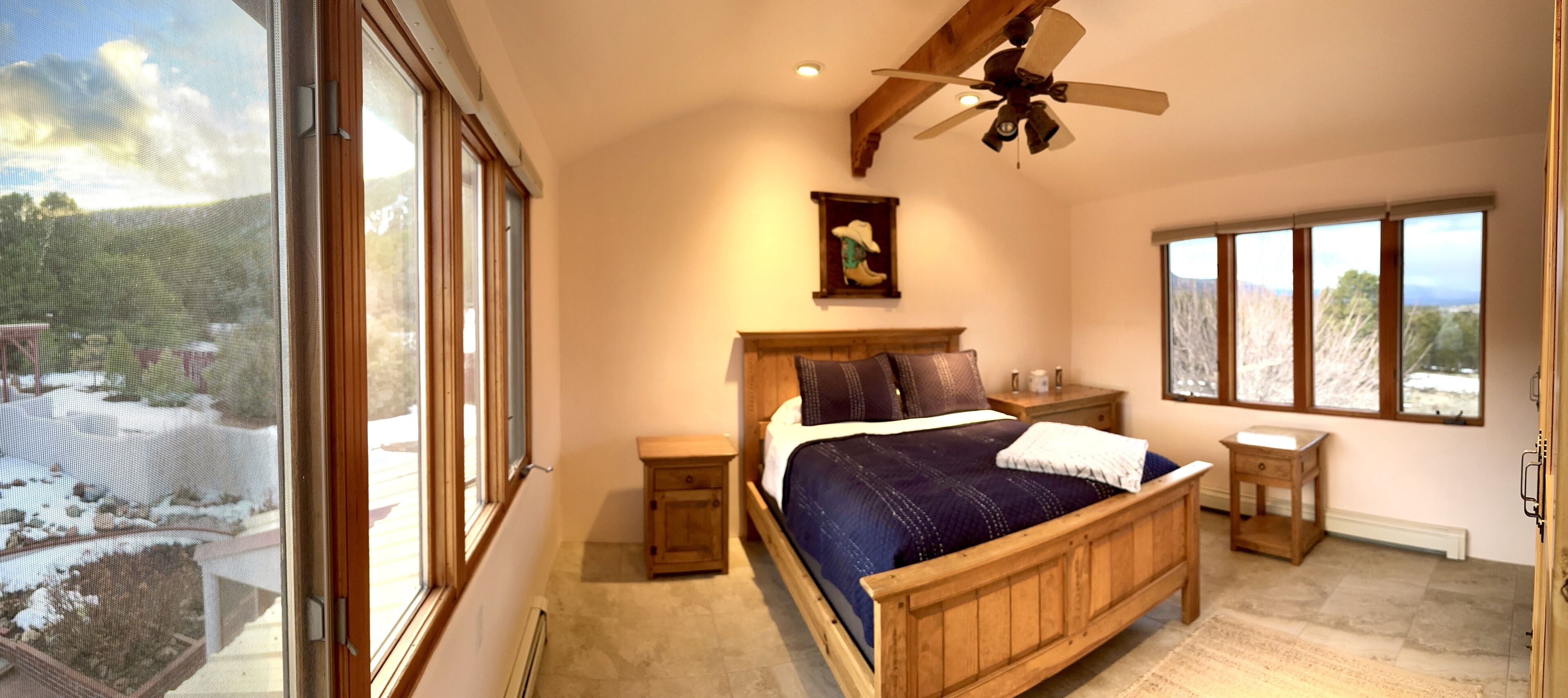 A bedroom with a large bed, wooden headboard, a ceiling fan, a nightstand, a bedside table, and a view of a snowy landscape through large windows.