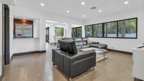 A modern living room with a black leather sofa, a kitchen area with a stainless steel refrigerator, and large windows offering a view of greenery. - 4BR Steps to Beach Home Private Pool, Hot Tub, BBQ (Fort Lauderdale)