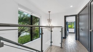 A modern interior with a glass railing, a chandelier, and a view of greenery outside. - 4BR Steps to Beach Home Private Pool, Hot Tub, BBQ (Fort Lauderdale)