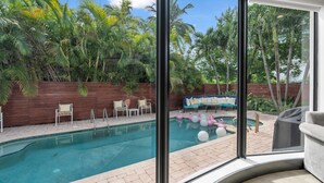 A pool with a wooden deck and lounge chairs, surrounded by lush greenery and palm trees. - 4BR Steps to Beach Home Private Pool, Hot Tub, BBQ (Fort Lauderdale)