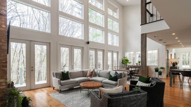 A spacious living room with a large sectional sofa, a round coffee table, and a fireplace. There are multiple windows providing natural light and views of the outdoors.