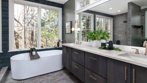 A modern bathroom with a freestanding bathtub, dark cabinetry, a large mirror, and a window with a view of trees.