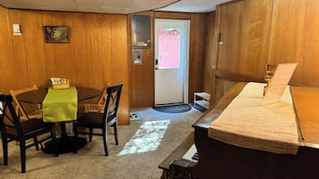 Una habitación con un piano, una mesa de comedor con sillas, un plato para perro pequeño y una puerta que da al exterior.