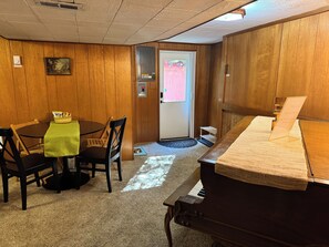 A room with a piano, a dining table with chairs, a small dog bowl, and a door leading outside.