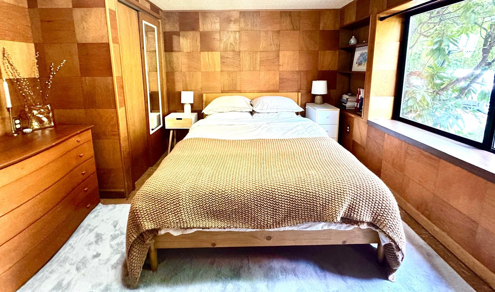 Un dormitorio con cama de madera, lámparas de noche, cómoda y ventana con vistas a la vegetación.