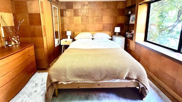 Un dormitorio con cama de madera, lámparas de noche, cómoda y ventana con vistas a la vegetación.