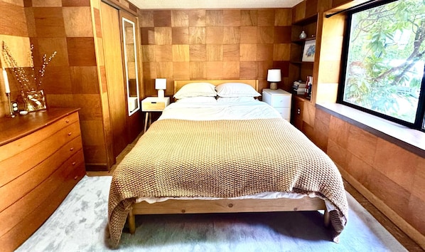 A bedroom with a wooden bed, bedside lamps, a dresser, and a window with a view of greenery.