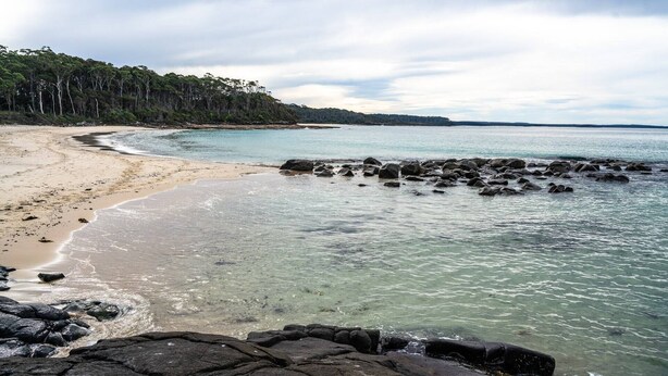 Ein Strand mit klarem Wasser, sandigem Ufer und einem bewaldeten Hügel im Hintergrund.