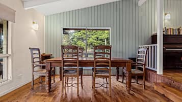 A dining area with a wooden table and chairs, a piano, and a view of greenery outside.