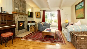 Ein gemütliches Wohnzimmer mit Kamin aus Stein, einem Klavier, einer Couch, einem Couchtisch und einem Korbsessel.