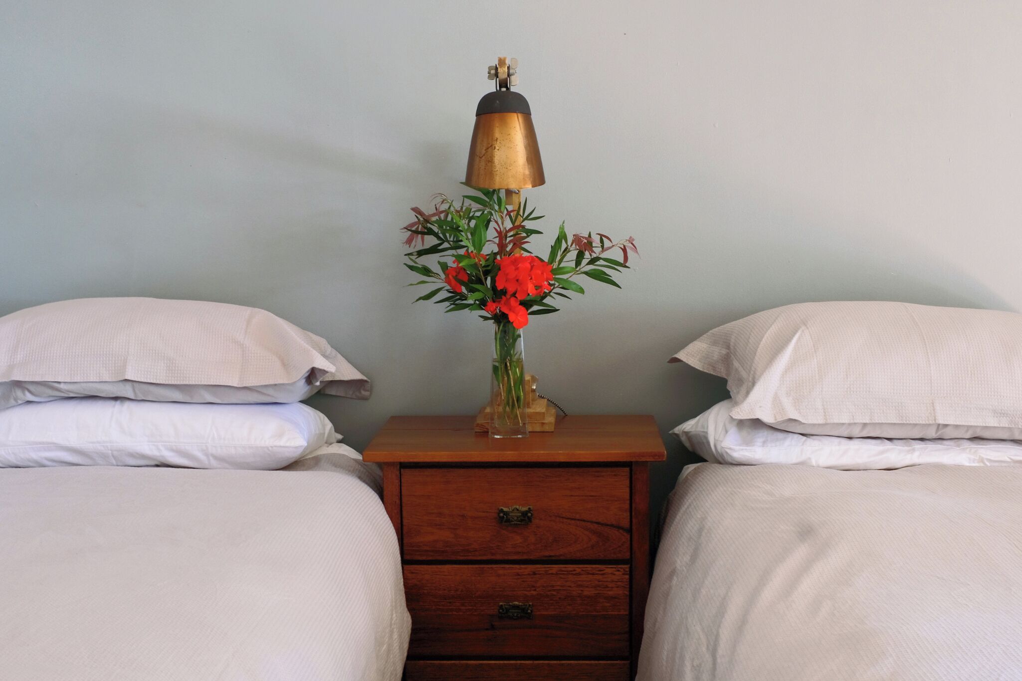 Une chambre avec deux lits, une table de chevet avec une lampe et un vase de fleurs, et un tiroir en bois.