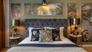 Une chambre à coucher avec une tête de lit capitonnée, un lit agrémenté de plusieurs coussins, deux tables de chevet avec des lampes et des œuvres d’art encadrées représentant un feuillage tropical.