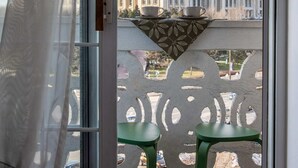 Un balcon donnant sur un grand immeuble, deux chaises vertes et une table avec deux tasses.