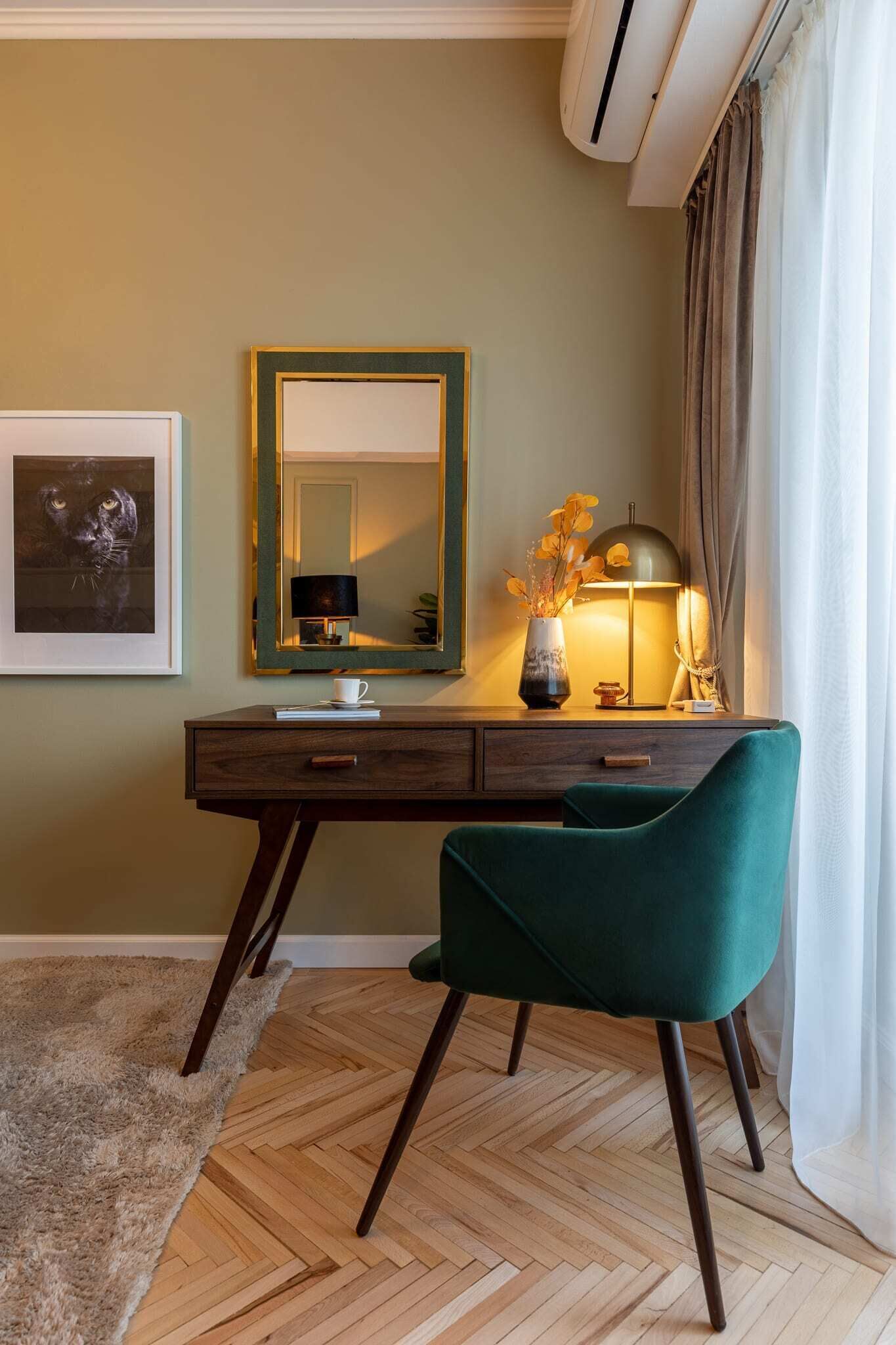 A room with a wooden herringbone floor, a desk with a mirror, a teal chair, and a lamp.