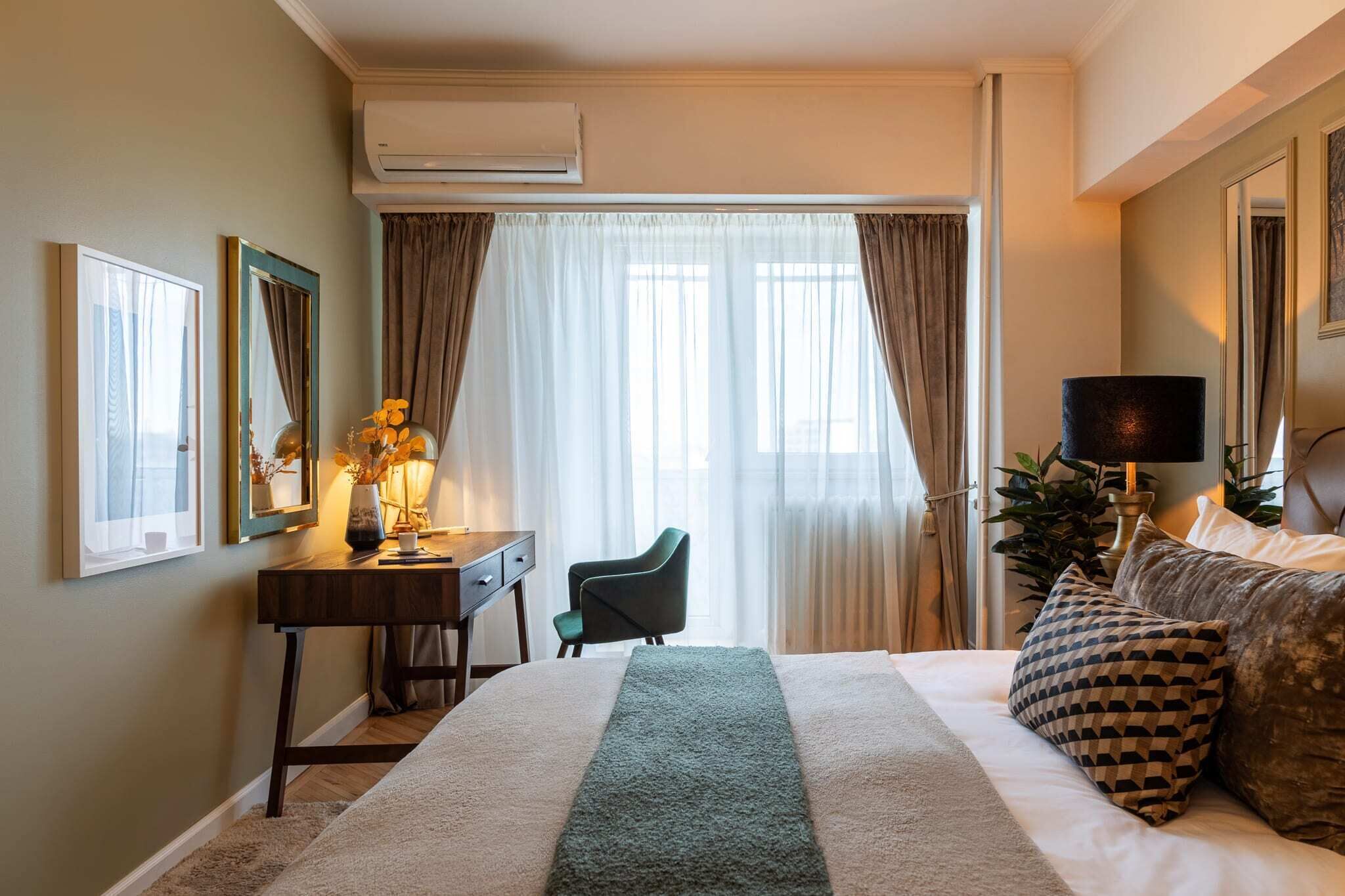 A hotel room with a bed, a desk, a chair, a lamp, and a plant.