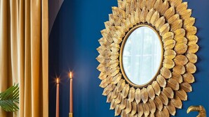 Une pièce avec un mur bleu, un miroir orné d’un motif de feuilles dorées, deux bougies allumées, une sculpture d’oiseau décorative et des meubles en osier.