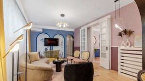 Un salon moderne avec un canapé incurvé, un fauteuil noir et une table basse. Un téléviseur est fixé au mur et une bibliothèque contient des objets décoratifs.