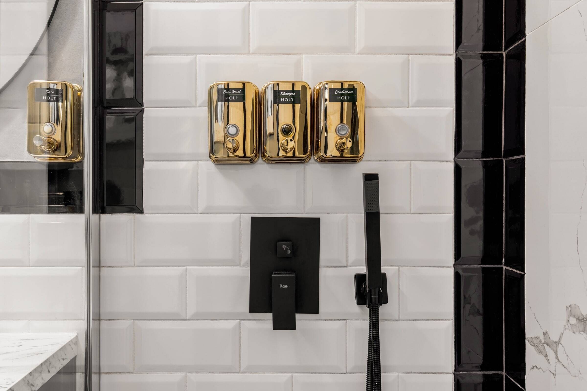 A bathroom with three gold-colored shower heads labeled "Baby Shower," "Shampoo," and "Chillwater" by HOLT, a black faucet, and a black handheld shower attachment.